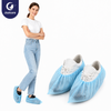 A smiling woman in a white T-shirt and blue jeans wearing Justcare disposable blue PP shoe covers, standing beside a pair of white casual shoes with the same shoe covers, on a white background — demonstrating easy wear, secure fit and everyday comfort.