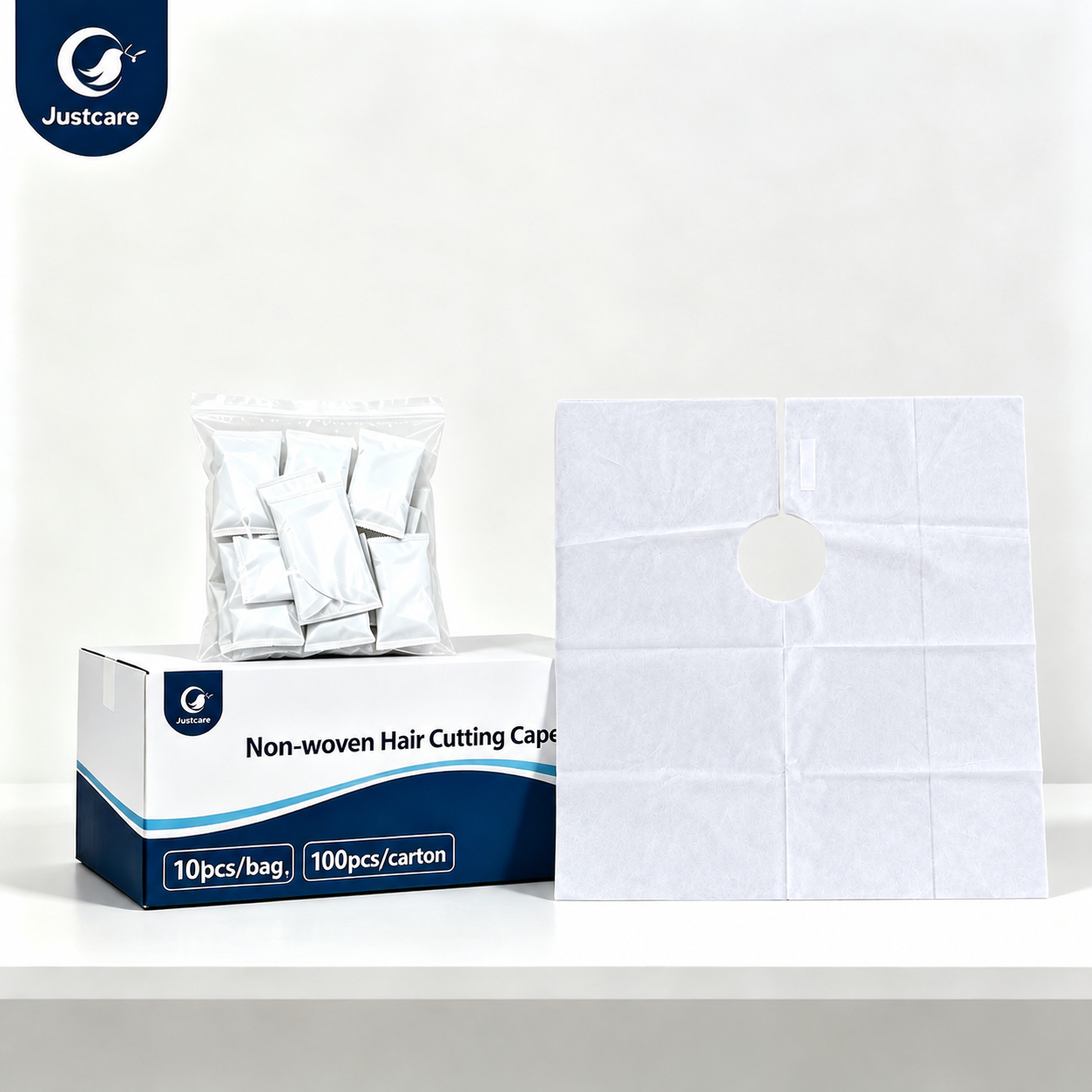 Justcare disposable non-woven hair cutting cape product shot. White cape, individually packed samples and outer carton on white table. 10pcs per bag, 100pcs per carton, clean plain white background